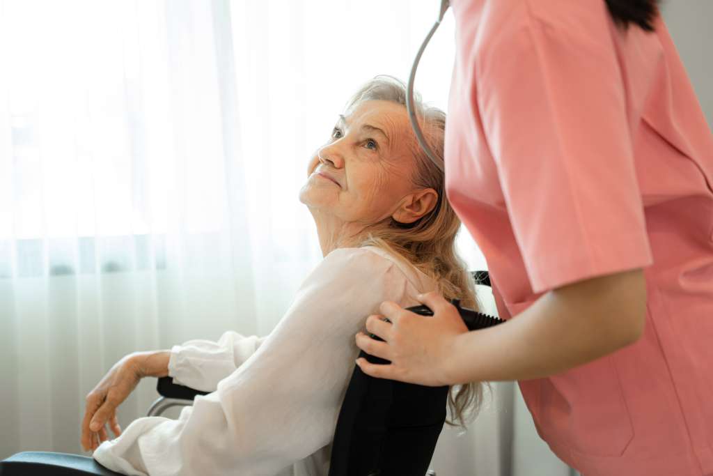 A nurse in pink scrubs pushing the wheelchair of an elderly woman in a memory care community.